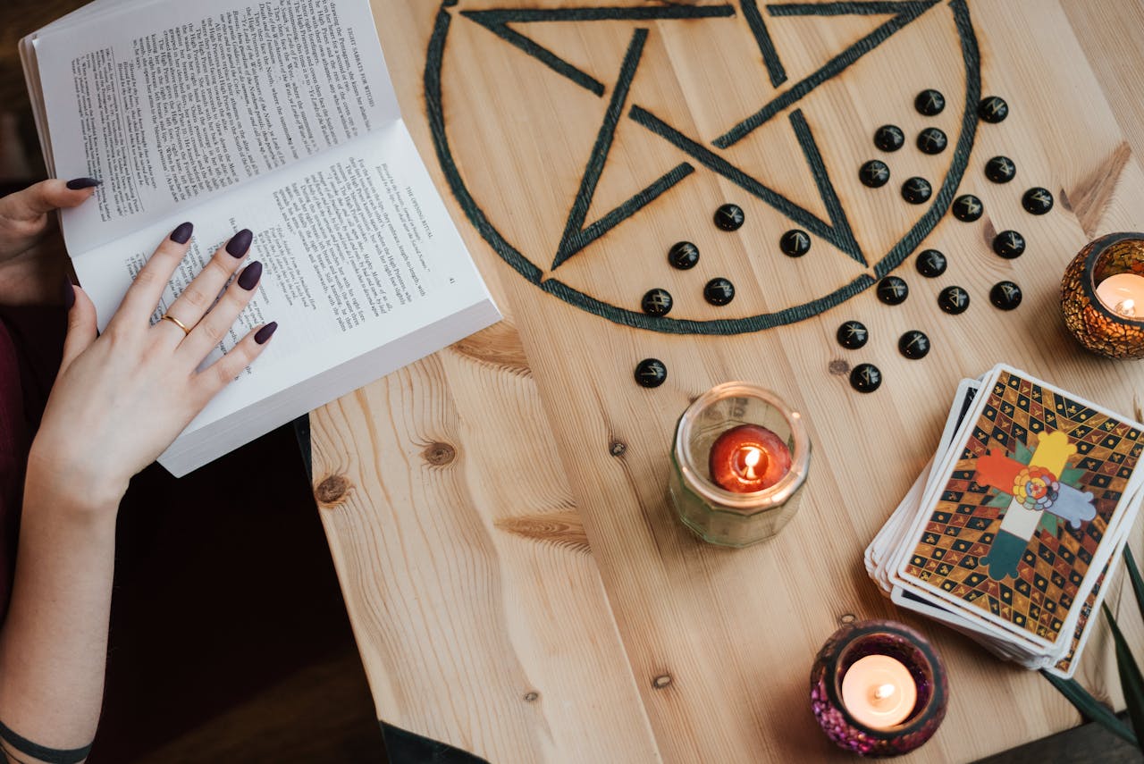 From above of crop unrecognizable fortune teller reading textbook at table with tarot cards and candles during divination process